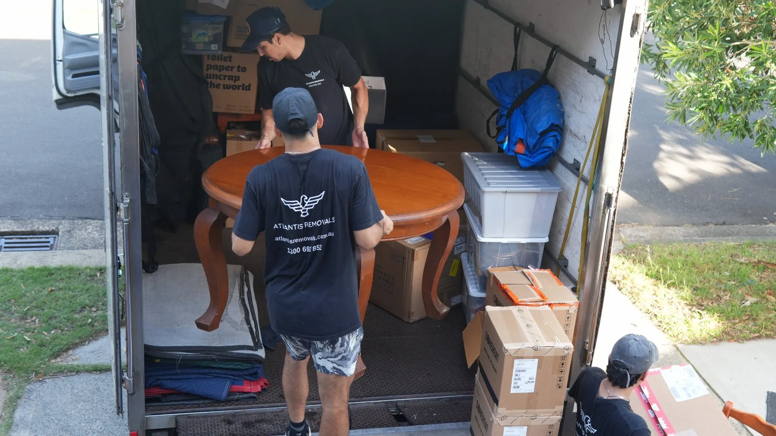 Atlantis Removals team members carefully handling a large wooden dining table and moving boxes inside a truck for a local home removal in Newcastle.