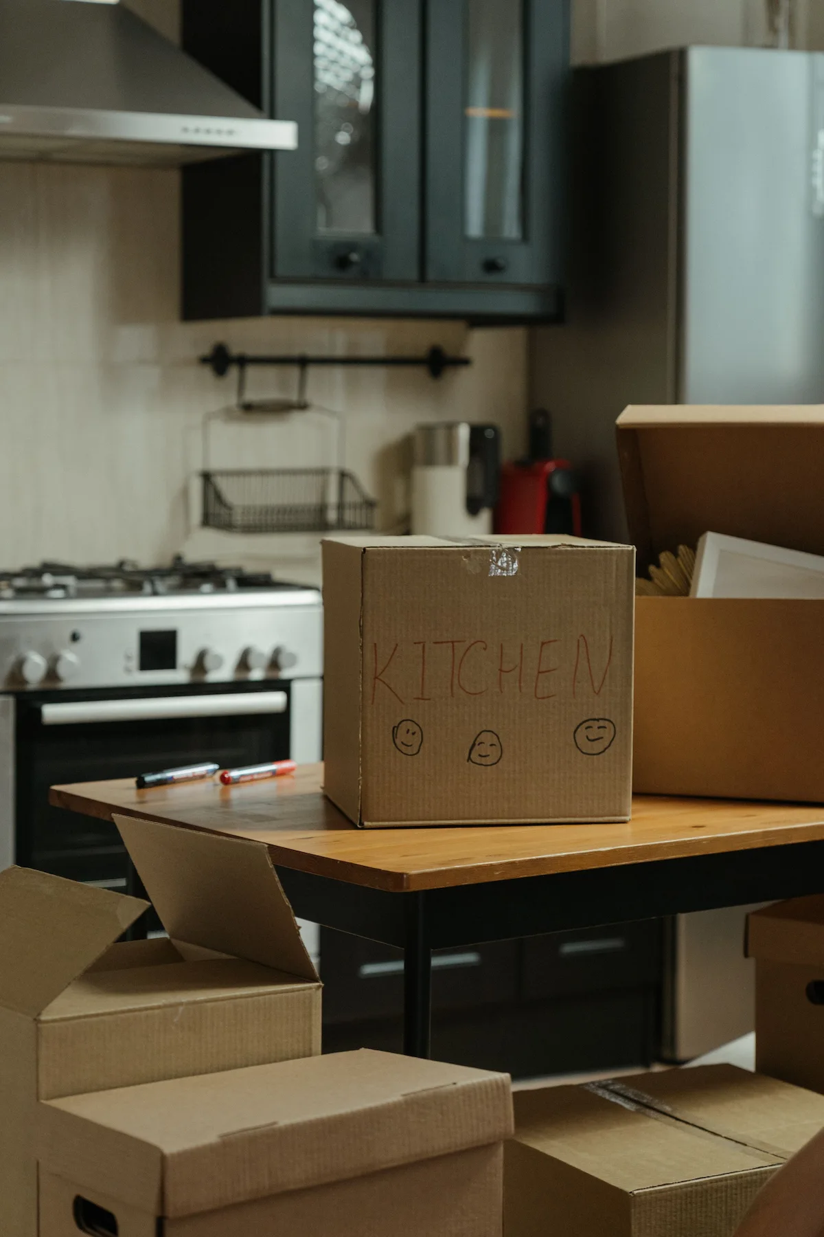 A neatly packed moving box handwritten with the word 'Kitchen' sitting on a wooden table surrounded by other boxes in a modern home.